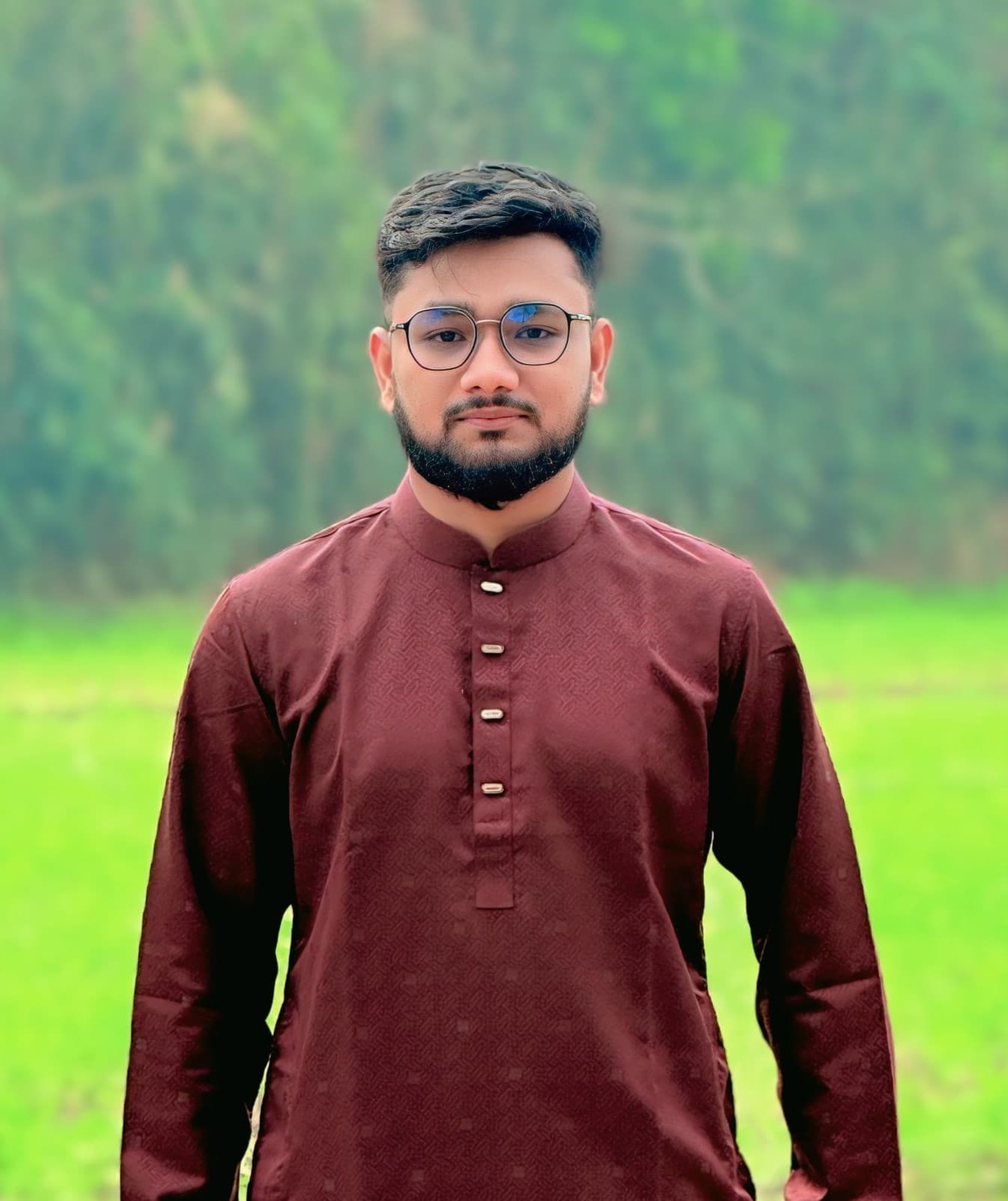 Imran Hossen in a maroon panjabi, standing outdoors in front of a green, wooded hillside.