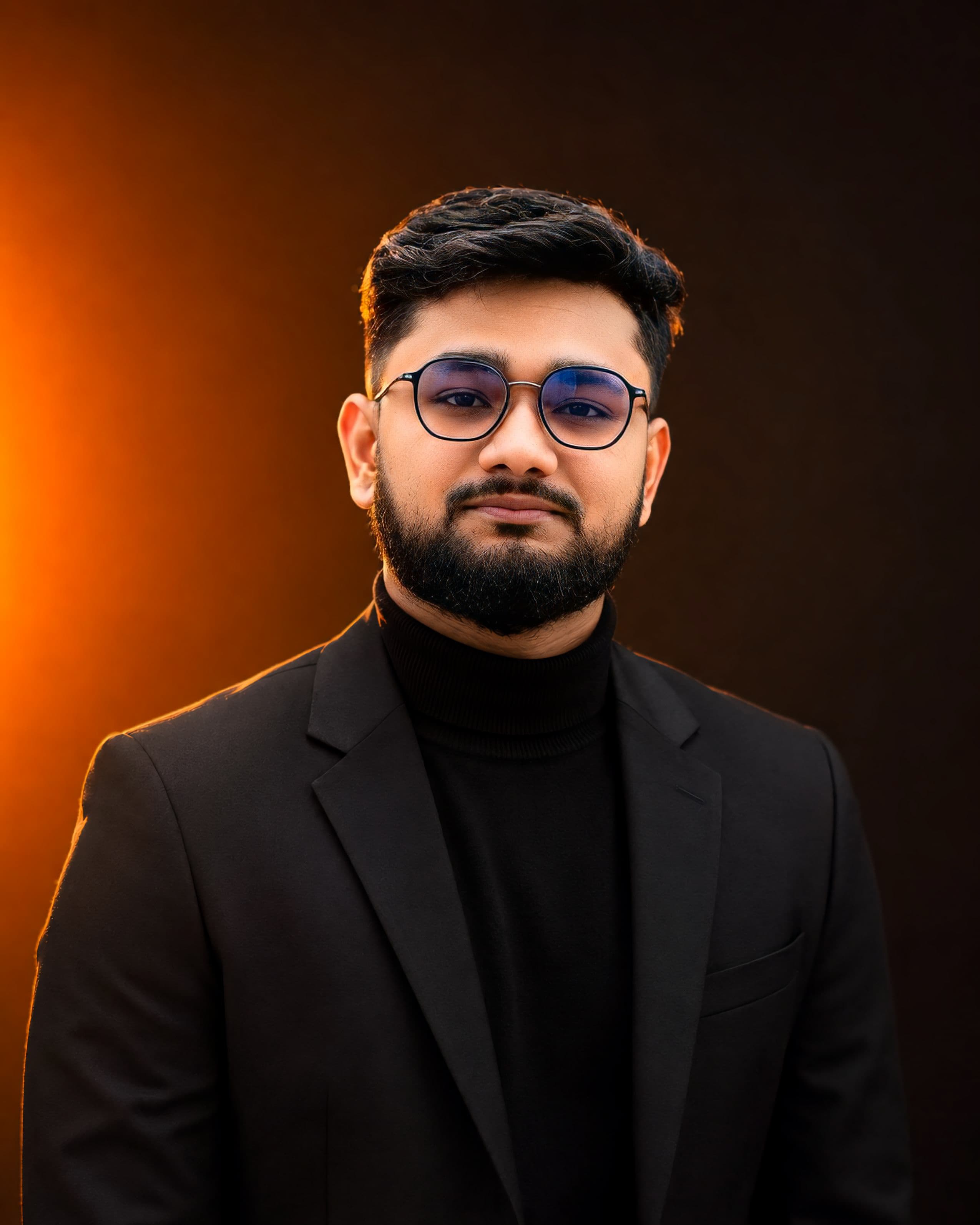 Imran Hossen in a black blazer and turtleneck, lit from the left by warm amber light against a dark background.